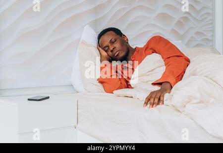 Jeune homme afro-américain portant une chemise orange dormant paisiblement dans un lit avec des draps blancs tout en tenant la main près du visage, avec smartphone reposant sur une table de nuit blanche Banque D'Images