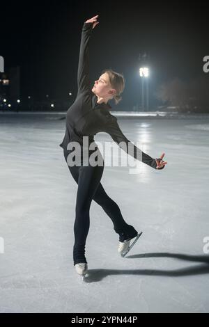 Une jeune femme en noir athlétique porte gracieusement des patins à glace sur une patinoire extérieure éclairée la nuit, entourée d'un éclairage doux. Banque D'Images