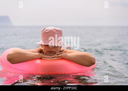 Une femme flotte sur un tube gonflable rose dans l'océan. Elle porte un chapeau rose et elle profite de l'eau. Banque D'Images