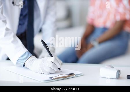 Gros plan d'un docteur méconnaissable portant des gants écrivant sur le presse-papiers consultation à la clinique avec le patient en arrière-plan flou, espace de copie Banque D'Images
