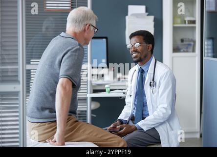 Portrait vue de côté d'un amical médecin afro-américain portant des lunettes et souriant tout en écoutant un patient senior expliquer des problèmes de santé en clinique Banque D'Images