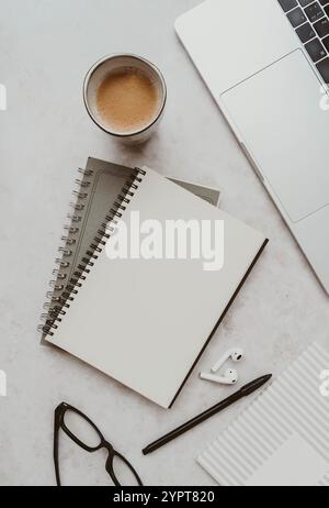 Espace de travail moderne avec ordinateur portable, cahiers en spirale, papier blanc, stylo noir, lunettes, tasse à café, et écouteurs sans fil sur le bureau blanc - professionnel minimal Banque D'Images