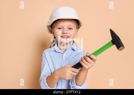 Petite fille en casque dur avec marteau sur fond beige. Rêver d'un futur métier Banque D'Images