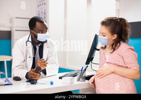 Médecin afro-américain explique les suppléments prénataux à la femme caucasienne pendant son bilan de grossesse. La mère attend écoute attentivement le médecin masculin tenant bouteille de pilules dans le cabinet médical. Banque D'Images