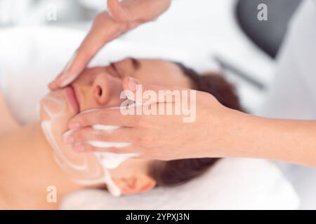 Crop anonyme cosmétologue massant le visage de la femme au salon de beauté. D'en haut de la cliente féminine détendue se fait dorloter dans le spa par esthéticienne. Banque D'Images