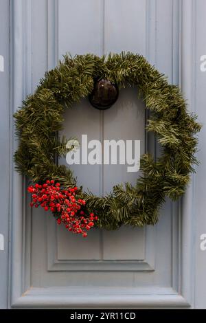 Une couronne de Noël classique à feuilles persistantes ornée de baies rouges éclatantes est accrochée à une porte bleu clair, évoquant l'esprit et la chaleur des fêtes. Banque D'Images