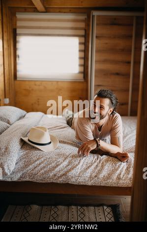 Beau jeune homme souriant assis sur le lit dans une maison en bois. Banque D'Images