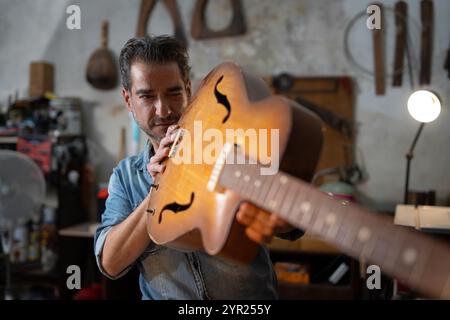 Fabricant professionnel de guitare inspectant les détails de guitare acoustique dans un atelier traditionnel. Artisan hispanique examinant l'instrument vintage montrant expertis Banque D'Images