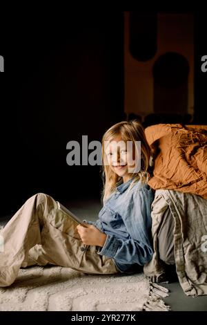 Portrait de garçon souriant assis avec livre près du lit sur le sol dans la chambre à la maison Banque D'Images