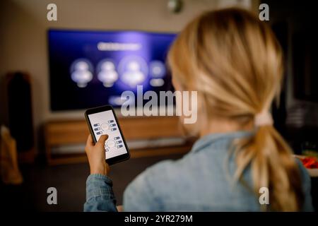 Vue arrière du garçon utilisant le téléphone intelligent comme télécommande pour regarder la télévision à la maison Banque D'Images