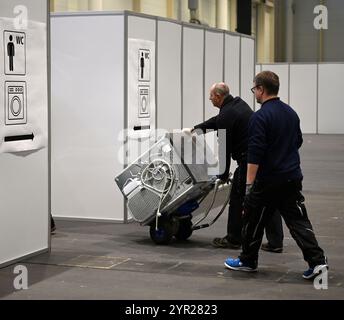 02 décembre 2024, Hambourg : deux ouvriers poussent une machine à laver dans les installations de lavage lors d'un rendez-vous avec les médias dans le logement des réfugiés dans le hall d'exposition B5 sur le terrain d'exposition. À partir du 3 décembre, le Hall B5 deviendra à nouveau un hébergement d’urgence pour les réfugiés. L'occupation est prévue pour jusqu'à 300 personnes, avec une capacité d'urgence totale de 476 places dans les compartiments. Le logement sera géré par l’entreprise sociale municipale Fördern & Wohnen. Photo : Niklas Graeber/dpa Banque D'Images