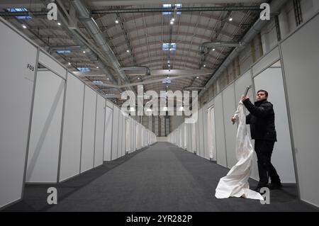 02 décembre 2024, Hambourg : un homme pose un rideau pour l'entrée d'une cabane lors de l'événement médiatique dans le logement des réfugiés dans le Hall B5 sur le terrain d'exposition. À partir du 3 décembre, le Hall B5 deviendra à nouveau un hébergement d’urgence pour les réfugiés. L'occupation est prévue pour jusqu'à 300 personnes, avec une capacité d'urgence totale de 476 places dans les compartiments. Le logement sera géré par l’entreprise sociale municipale Fördern & Wohnen. Photo : Marcus Brandt/dpa Banque D'Images