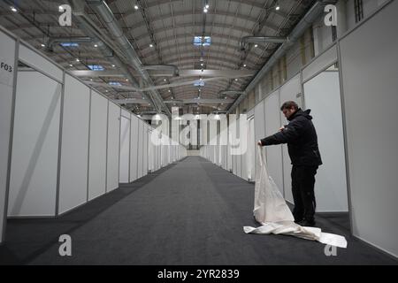 02 décembre 2024, Hambourg : un homme pose un rideau pour l'entrée d'une cabane lors de l'événement médiatique dans le logement des réfugiés dans le Hall B5 sur le terrain d'exposition. À partir du 3 décembre, le Hall B5 deviendra à nouveau un hébergement d’urgence pour les réfugiés. L'occupation est prévue pour jusqu'à 300 personnes, avec une capacité d'urgence totale de 476 places dans les compartiments. Le logement sera géré par l’entreprise sociale municipale Fördern & Wohnen. Photo : Marcus Brandt/dpa Banque D'Images