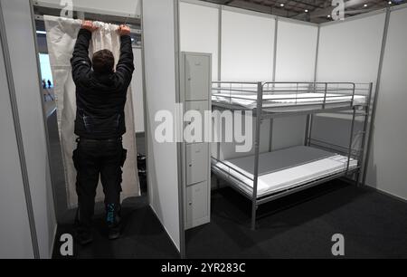 02 décembre 2024, Hambourg : un homme attache un rideau à l'entrée d'une cabine d'hébergement temporaire lors de la manifestation médiatique dans le logement des réfugiés du Hall B5 sur le terrain d'exposition. À partir du 3 décembre, le Hall B5 deviendra à nouveau un hébergement d’urgence pour les réfugiés. L'occupation est prévue pour jusqu'à 300 personnes, avec une capacité d'urgence totale de 476 places dans les compartiments. Le logement sera géré par l’entreprise sociale municipale Fördern & Wohnen. Photo : Marcus Brandt/dpa Banque D'Images