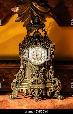 Une grande horloge ornée avec un cadre doré se trouve sur une table. L'horloge a un cadran et des aiguilles dorés, et les chiffres sont noirs. L'horloge est entourée d'un Banque D'Images