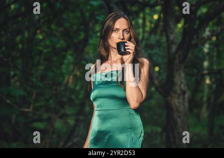 Femme adulte rêveuse avec du café dans la forêt verte en été. Grands yeux verts, cheveux longs, robe verte Banque D'Images