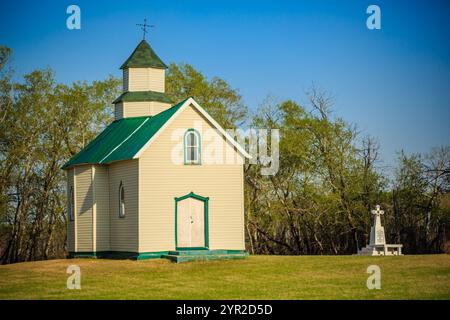 Une petite église blanche avec un toit vert se trouve dans un champ herbeux. L'église est entourée d'arbres et une croix est placée devant elle. La scène est pe Banque D'Images