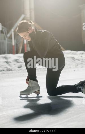 Une jeune femme en noir athlétique porte gracieusement des patins à glace sur une patinoire extérieure éclairée la nuit, entourée d'un éclairage doux. Banque D'Images