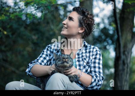 Jeune femme est assise dans une forêt, tenant son chat tabby, levant les yeux avec un sourire, profitant de la nature. Ils partagent un moment d'amour et de bonheur, entourés de b. Banque D'Images