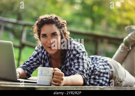 Une jeune femme d'une vingtaine d'années travaille à distance dans un parc paisible, en utilisant son ordinateur portable et en dégustant une boisson chaude Banque D'Images