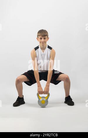 Adolescent de quinze ans accroupissant avec une kettlebell, photographié en studio sur fond blanc Banque D'Images