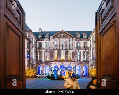 Strasbourg, France - 29 novembre 2024 : la cour de l'hôtel de ville de Strasbourg décorée pour Noël avec des lumières festives, des arbres et des visiteurs passant par une grande porte en bois au crépuscule Banque D'Images