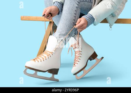 Jeune femme en vêtements d'hiver avec des patins à glace attachant des lacets sur le banc sur fond bleu, gros plan Banque D'Images