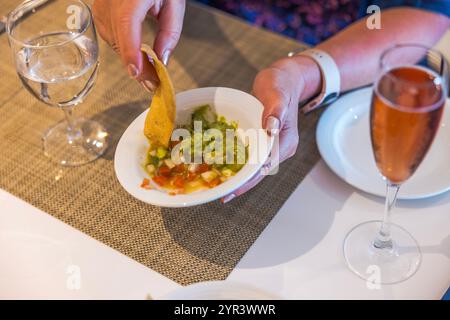 Main tenant la puce de tortilla trempant dans le guacamole frais et la salsa dans un bol blanc entouré de verres à vin et à eau. Mexique. Banque D'Images