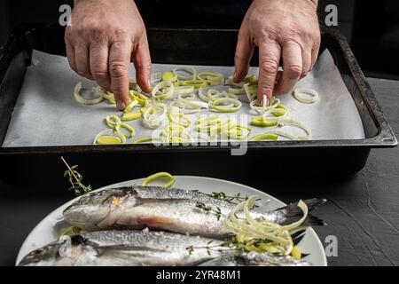 Les mains du chef étalent les poireaux sur du papier cuisson sur la protvina et le poisson dorado sur une assiette blanche, prêts à être cuits sur un fond noir. Le Banque D'Images