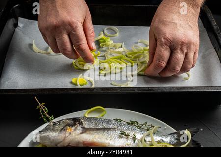 Les mains du chef étalent les poireaux sur du papier cuisson sur la protvina et le poisson dorado sur une assiette blanche, prêts à être cuits sur un fond noir. Le Banque D'Images