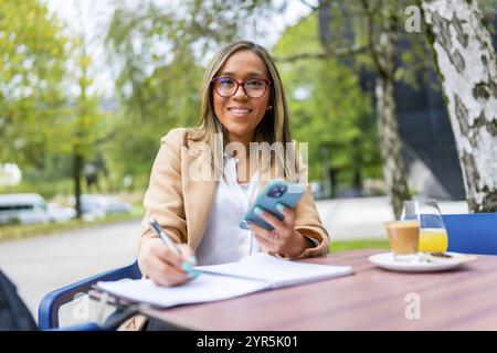 Femme latine utilisant le téléphone tout en écrivant des notes buvant du café assis sur une terrasse tranquille d'une cafétéria dans la ville Banque D'Images