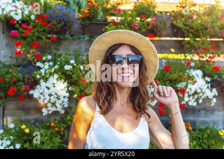 Une femme portant un chapeau de paille et des lunettes de soleil sourit devant un mur de fleurs. Les fleurs sont de différentes couleurs et sont disposées de manière à ce que Banque D'Images