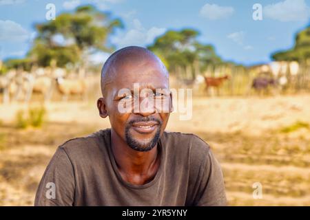 portrait de l'homme africain du village à la ferme en face de l'enclos à bétail dans une journée ensoleillée, moutons et chèvres dans les arrière-plans Banque D'Images