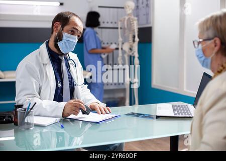 Femme retraitée attend sa prescription alors que le jeune médecin remplit les documents de santé en utilisant un tampon à l'hôpital. Médecin masculin estampillant sur le presse-papiers pendant que la patiente retraitée attend au bureau dans la clinique. Banque D'Images