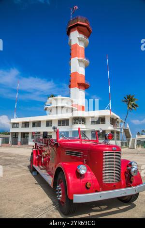 Un camion de pompiers rouge est garé devant un phare. La scène est calme et paisible, avec le ciel bleu et le phare en arrière-plan Banque D'Images