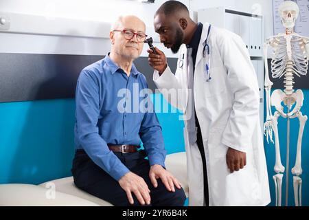 Homme blanc retraité assis sur le lit d'hôpital alors que le médecin afro-américain inspecte soigneusement son oreille pour l'infection. Médecin noir utilisant l'otoscope sur le sujet sénior au cours de l'examen médical. Banque D'Images