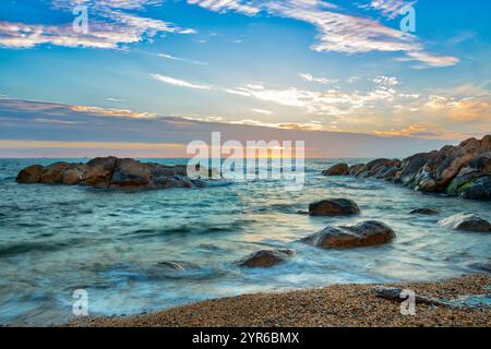 La lumière douce du soleil baigne une scène océanique tranquille, mettant en évidence les rochers et une plage de sable au premier plan, créant un landsca côtier serein et pittoresque Banque D'Images