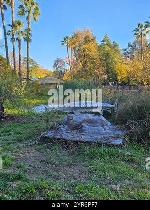 Sculpture d'esturgeon sur une base rocheuse entourée de verdure et de palmiers dans un parc naturel sous un ciel bleu clair. Banque D'Images