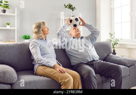 Couple senior retraité assis sur le canapé dans le salon à la maison et s'amuser avec le ballon de football. Banque D'Images
