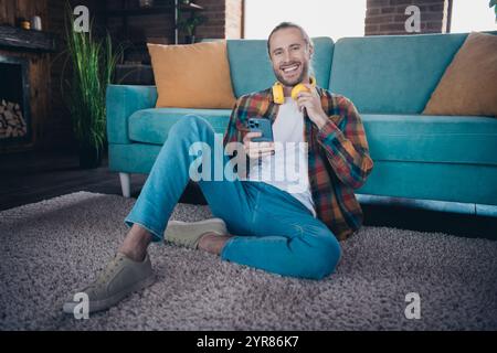 Jeune homme profitant d'un week-end de détente à l'intérieur avec écouteurs et smartphone, assis confortablement sur une élégante moquette Banque D'Images