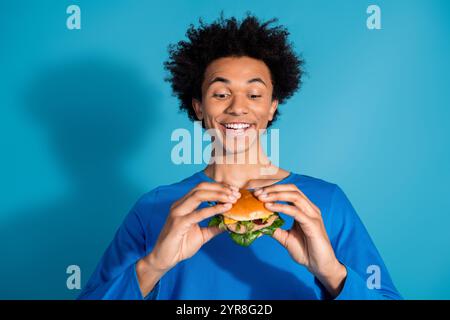 Portrait photo de beau jeune gars mange burger morsure habillé vêtement élégant isolé sur fond de couleur bleue Banque D'Images