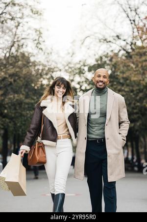 Un couple heureux se promène dans le parc en transportant des sacs à provisions par une agréable journée. Banque D'Images