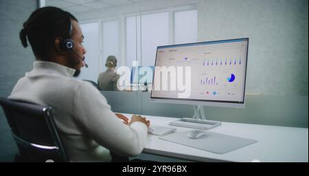 Professionnel afro-américain spécialiste financier ou gestionnaire de commerce électronique dans le casque consulte le client, travaille sur ordinateur. Écran PC avec données commerciales et diagrammes. Bureau moderne de la société E-business Banque D'Images