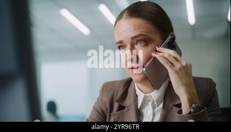 Femme caucasienne travaille dans un bureau avancé lumineux : spécialiste du marketing féminin stressé ou gestionnaire de clientèle parle agressivement au partenaire commercial ou au client à l'aide du téléphone, regarde l'écran du PC. E-business. Banque D'Images