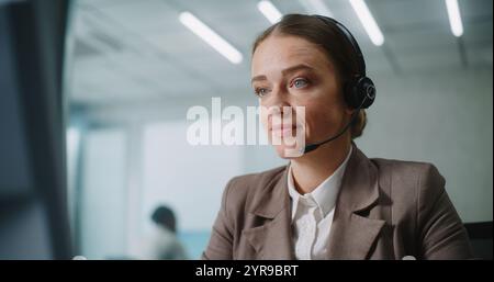 Femme caucasienne travaille dans un bureau moderne : opérateur de support technique féminin ou gestionnaire de clientèle parle sur casque avec le client, utilise un ordinateur, fournit un service client en ligne. E-business et E-commerce. Banque D'Images