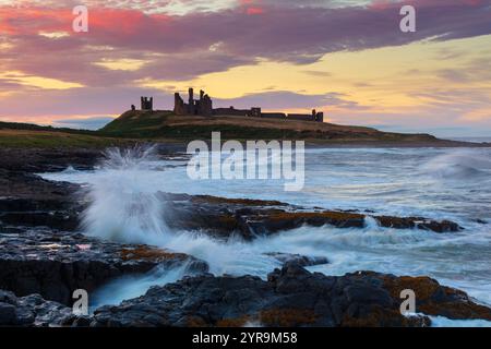 Vagues se brisant sur les rochers au coucher du soleil avec le château de Dunstanburgh au loin, Northumberland, Angleterre, Royaume-Uni. Banque D'Images