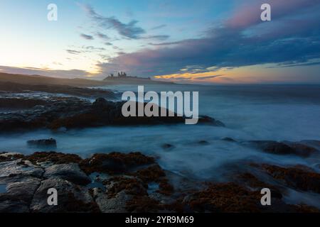 Vagues se brisant sur les rochers au coucher du soleil avec le château de Dunstanburgh au loin, Northumberland, Angleterre, Royaume-Uni. Banque D'Images