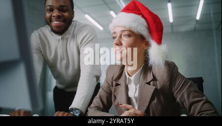 Spécialiste du marketing féminin à Santa Hat utilise un ordinateur, entre des données sur le clavier. Le chef d'équipe afro-américain aide ses collègues. Les hommes d'affaires travaillent dans un bureau moderne décoré pour Noël. E-commerce. Banque D'Images