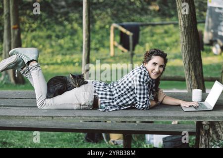 Jeune femme est allongée sur une table de pique-nique, travaillant sur son ordinateur portable avec son chat dormant sur le dos, profitant de l'air frais et du soleil dans un extérieur paisible Banque D'Images