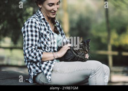 Une jeune femme assise sur un banc à l'extérieur dans un parc caresse un chat tabby allongé sur ses genoux, profitant d'un moment paisible de connexion avec la nature et lui Banque D'Images